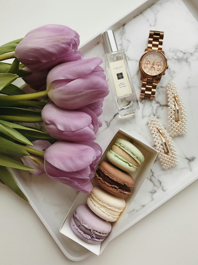 A stylish flat lay featuring purple tulips, macarons, a watch, and perfume on a marble tray.